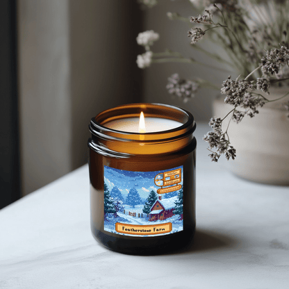 A lit Stardew Valley scented candle sits in an amber jar with a label depicting a snowy scene and the words "Featherstone Farm.