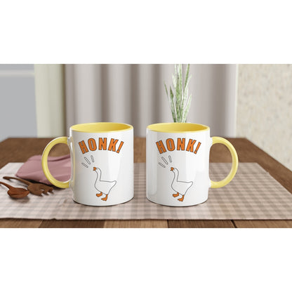 Two mugs with yellow rims and Untitled Goose Game illustration with orange 'Honk!' text on a table with a checkered tablecloth.