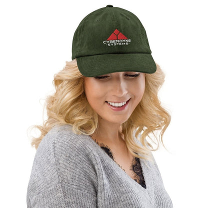 A person wearing a green corduroy baseball cap featuring the Cyberdyne Systems logo embroidered on the front.