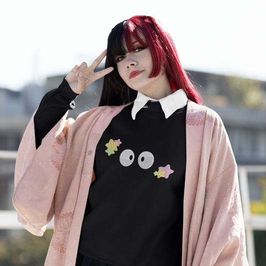 A person wearing a black Soot Sprites embroidered sweater inspired by Studio Ghibli poses outdoors with one hand raised in a peace sign.