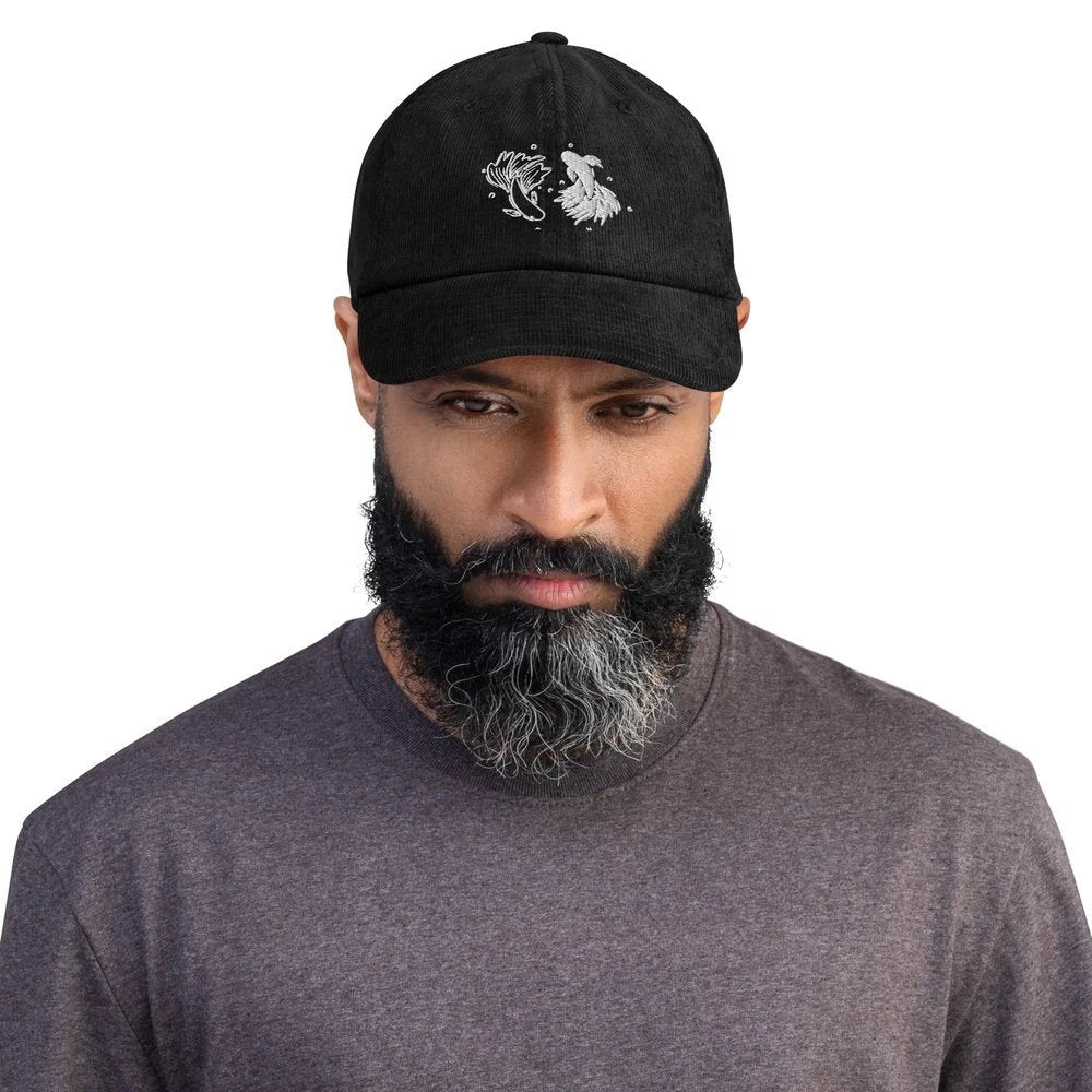 A person wearing a black Jujutsu Kaisen embroidered baseball cap featuring a betta fish design resembling a yin yang symbol.