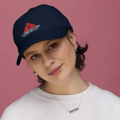 A person wears a dark blue baseball cap featuring the Cyberdyne Systems logo embroidered on the front.