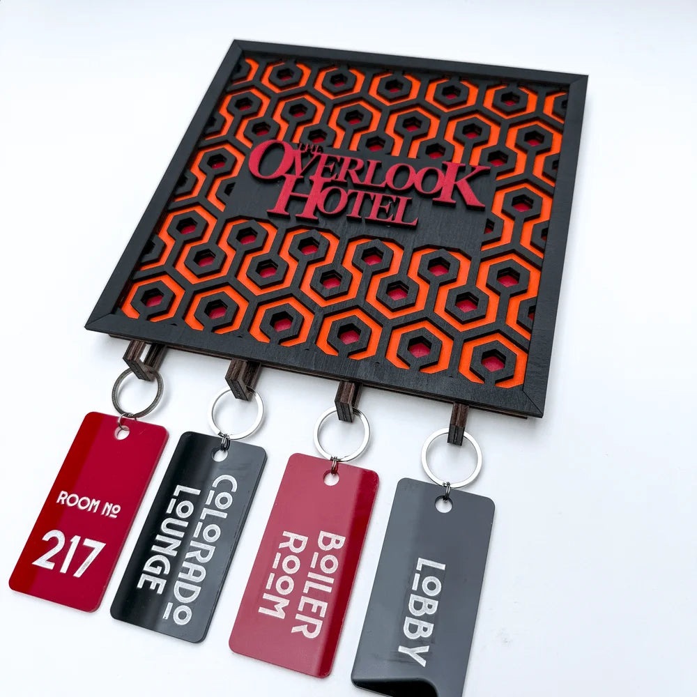 A wooden laser-cut key holder featuring a carpet pattern and "Overlook Hotel" lettering from The Shining is displayed with four room keychains labeled Room No. 17, Colorado Lounge, Boiler Room, and Lobby.