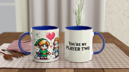 A white mug with a blue handle features pixel art of Link and Zelda from The Legend of Zelda, along with the phrase "You're My Player Two.