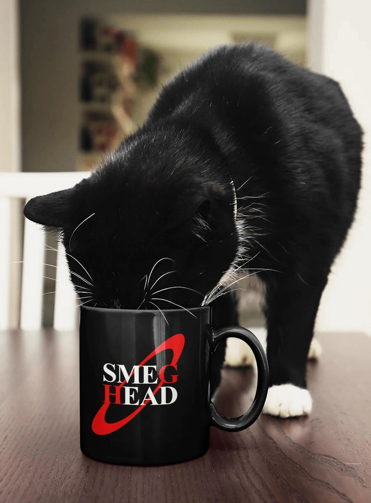 A black cat leans over a black Smeg Head mug sitting on a wooden table.