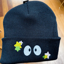 A black Soot Sprites beanie features embroidered eyes and yellow and green stars inspired by Ghibli's Susuwatari.