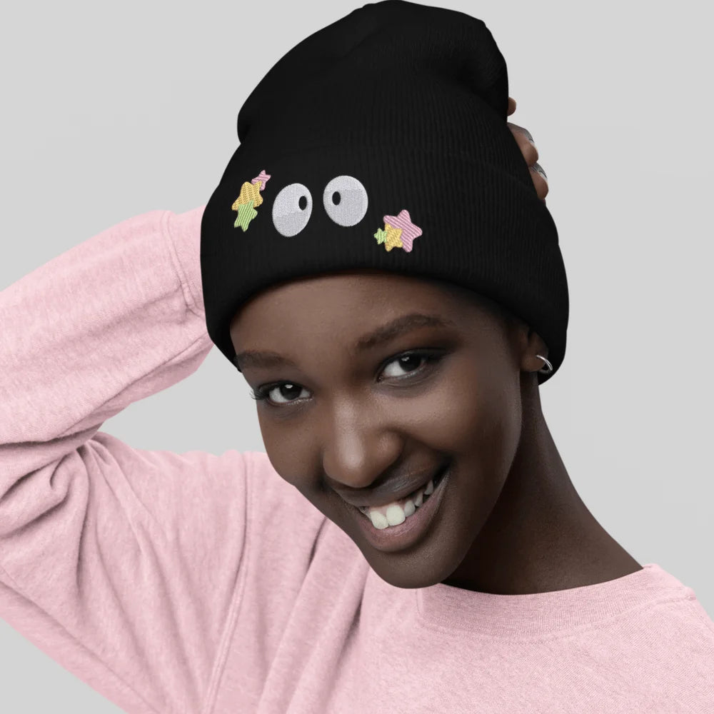 A person wearing a black Soot Sprites beanie featuring embroidered Susuwatari and star designs inspired by Ghibli.