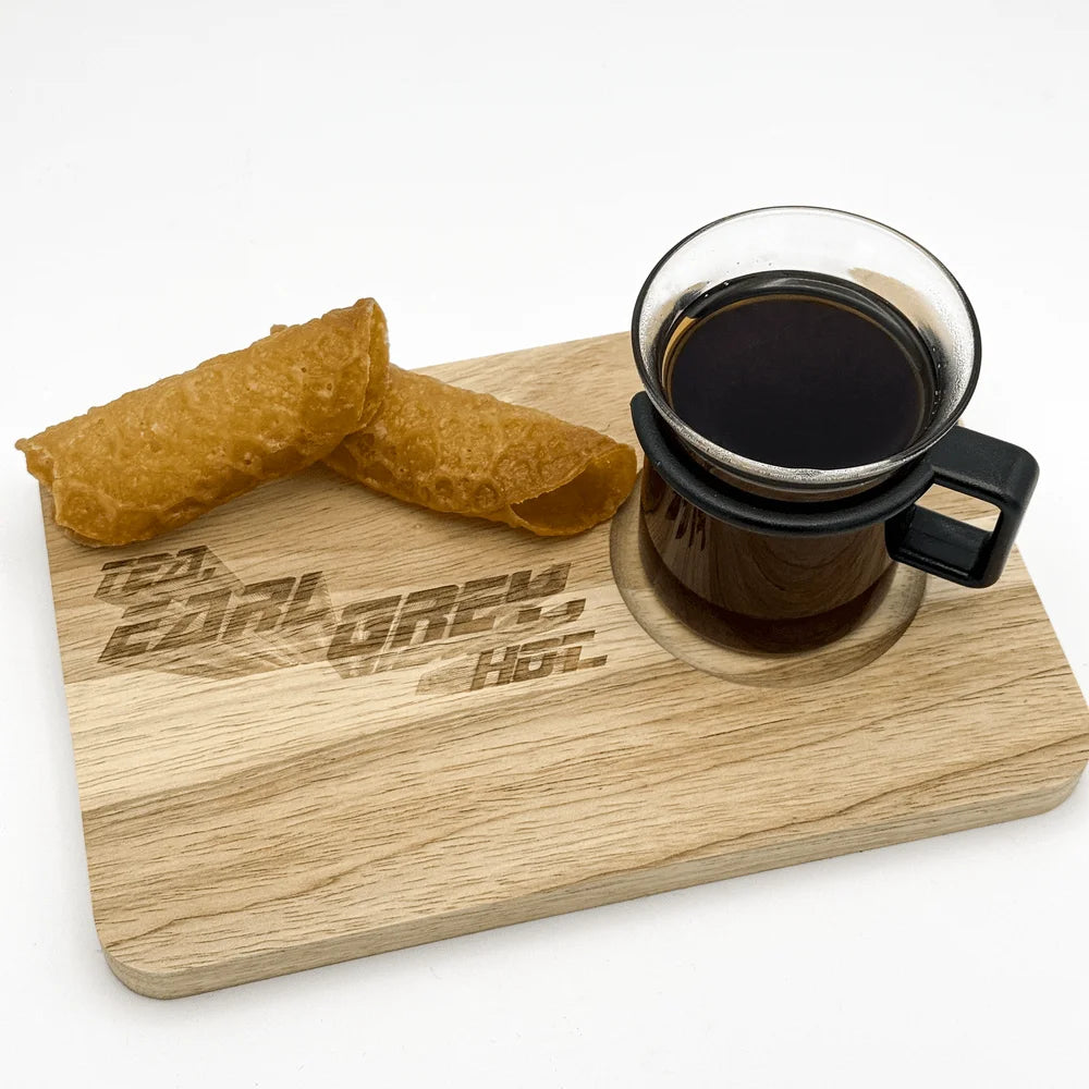 A wooden chopping board displays "Tea, Earl Grey, Hot" engraved lettering and holds a cup of dark liquid alongside a folded fortune cookie.