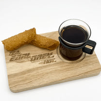 A wooden chopping board displays "Tea, Earl Grey, Hot" engraved lettering and holds a cup of dark liquid alongside a folded fortune cookie.