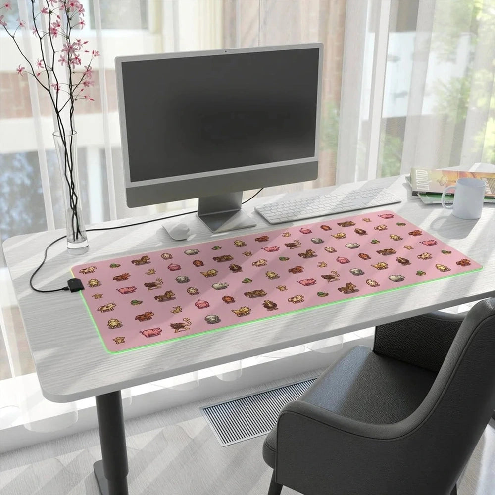 A pink Stardew Valley desk mat featuring pixel art of animals sits on a wooden desk next to a computer monitor and office supplies.