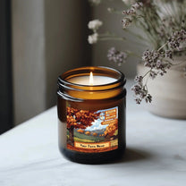 A Stardew Valley-themed amber glass candle burns on a marble surface, featuring an illustrated scene of a farm and personalized text at the bottom.