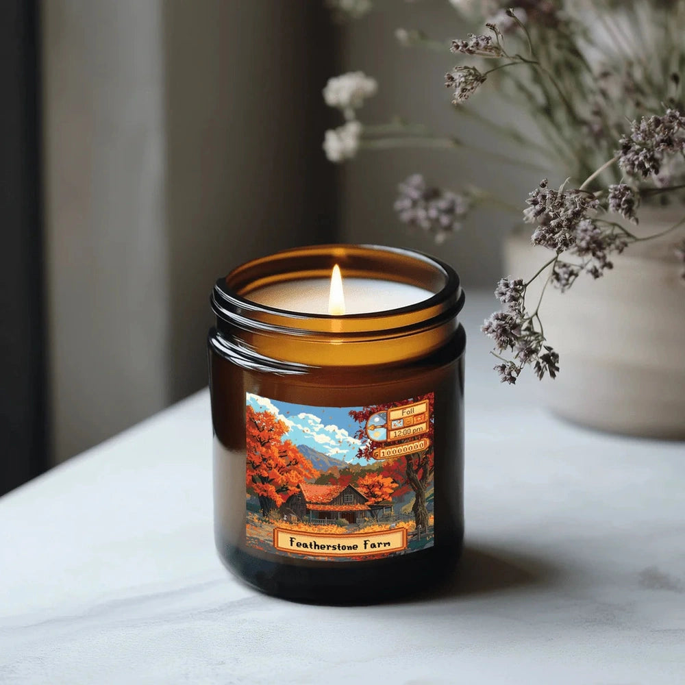 A lit Stardew Valley scented candle is displayed in an amber jar featuring a landscape illustration of Featherstone Farm and autumn foliage.