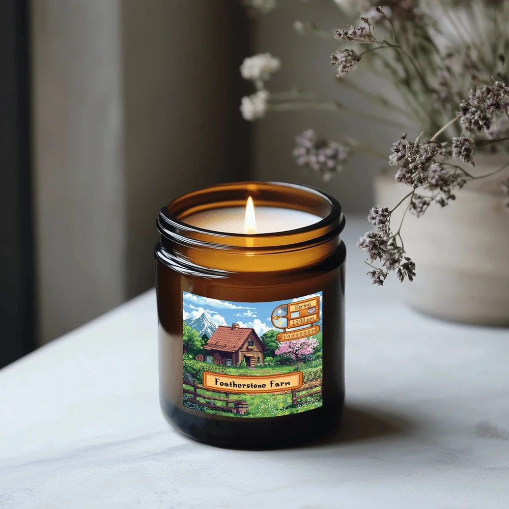 A lit Stardew Valley-themed candle in a brown jar sits on a white surface, displaying an illustration of Featherstone Farm and seasonal scent labels.
