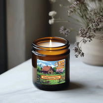 A lit Stardew Valley-themed candle in a brown jar sits on a white surface, displaying an illustration of Featherstone Farm and seasonal scent labels.