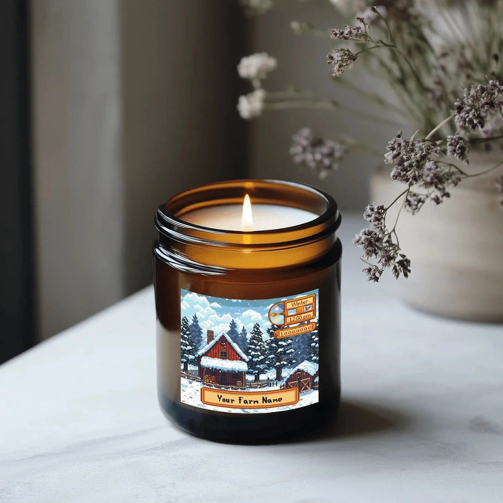 A lit Stardew Valley scented candle is displayed in an amber jar featuring a snowy farm scene and personalized text.