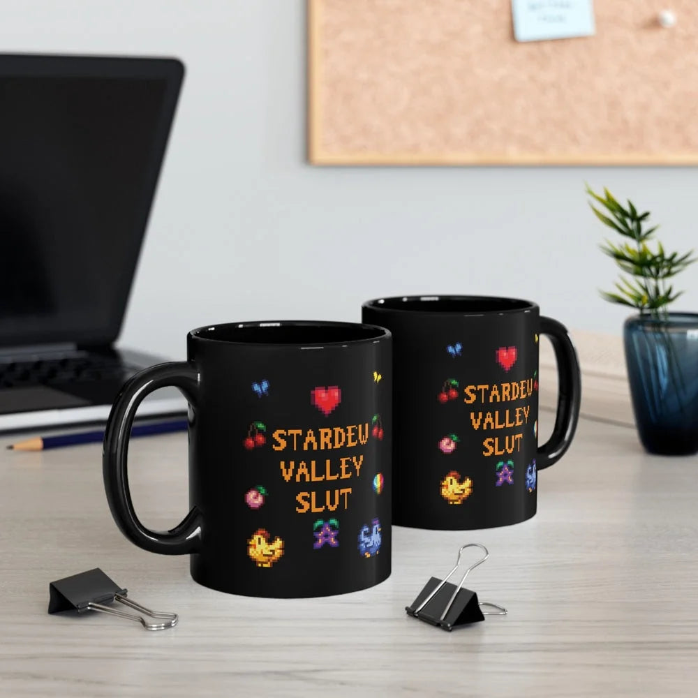 Two black mugs featuring pixel art graphics and the text Stardew Valley Slut sit on a desk next to a laptop and binder clips.