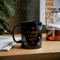 A black mug featuring pixel art graphics and the text Stardew Valley Slut sitting on a wooden table next to a glass teapot.