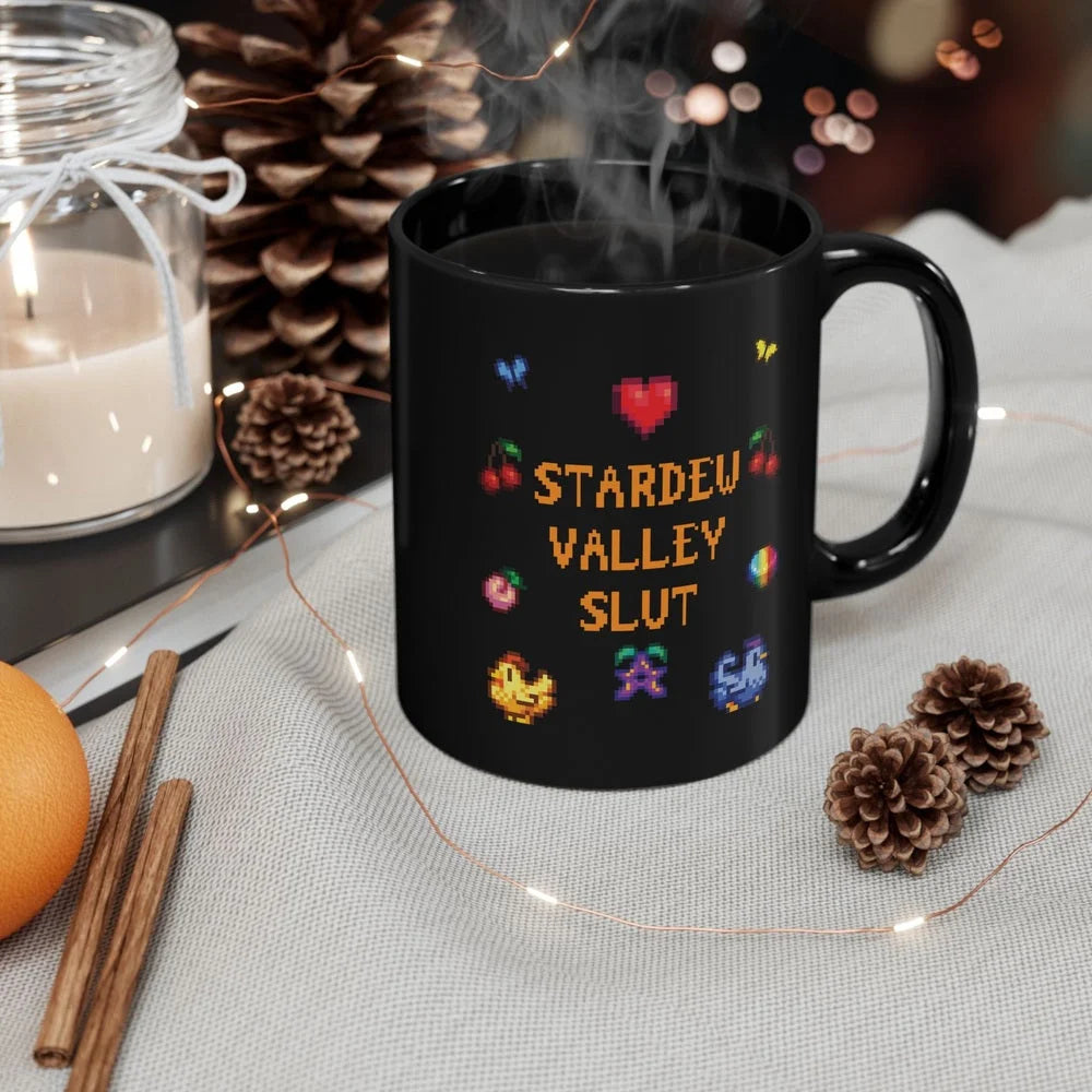 A black mug featuring pixel art graphics and the text Stardew Valley Slut sits on a cloth next to pinecones and a candle.