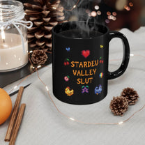 A black mug featuring pixel art graphics and the text Stardew Valley Slut sits on a cloth next to pinecones and a candle.