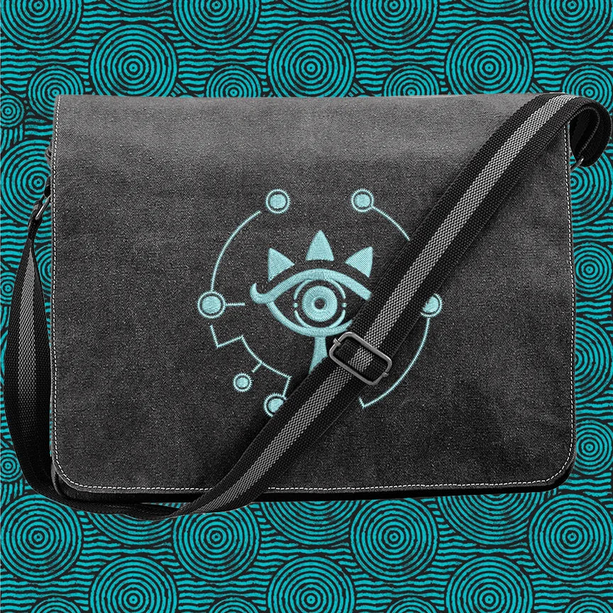 A black Zelda Sheikah messenger bag features glow-in-the-dark embroidery of a stylized eye symbol on the front flap and an adjustable shoulder strap.