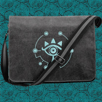 A black Zelda Sheikah messenger bag features glow-in-the-dark embroidery of a stylized eye symbol on the front flap and an adjustable shoulder strap.